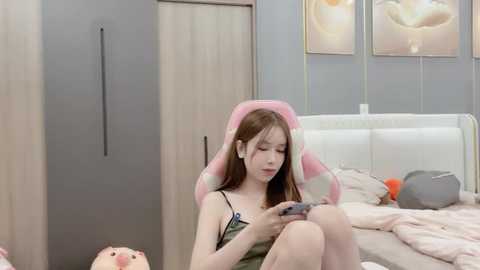 Media: Video of an Asian woman with long brown hair, wearing a green camisole, playing a video game on a pink gaming chair in a modern bedroom with light gray walls, white bed, and floral art.