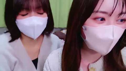 Media: A video shows two young Asian women with straight, dark hair and fair skin, wearing white surgical masks and light-colored blouses, seated indoors with a green wall background.