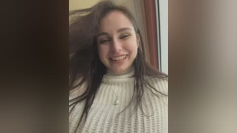 Media: Video of a smiling young woman with long brown hair, wearing a cream knitted turtleneck sweater, standing indoors next to a window with a beige wall in the background.