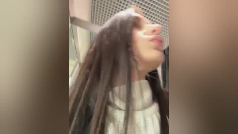 Media: Video of a young woman with long, straight, dark hair, wearing a white sweater, standing in a beige hallway with a grid-patterned wall and closed door.