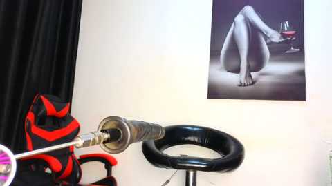 Media: Video of a minimalist studio featuring a black chair and a red and black backpack on a white wall with a serene, black-and-white nude video of a woman sitting on a chair.