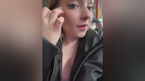 Media: Video of a young woman with fair skin, light brown hair, wearing a black leather jacket, adjusting her earring with her left hand, sitting in a public bus.