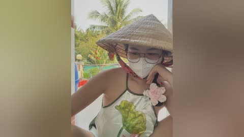 Media: Video of a young woman with light skin, wearing a traditional Vietnamese conical hat, glasses, and mask, sitting in a garden with a pond and palm trees. She has a white dress with a green leaf design and a pink flower in her hair.