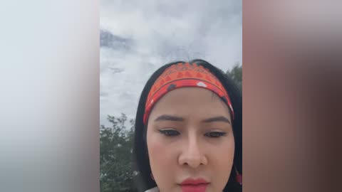 Media: Video of a young Asian woman with long black hair and a red headband, her expression neutral, in a blurred outdoor setting with cloudy sky and greenery.