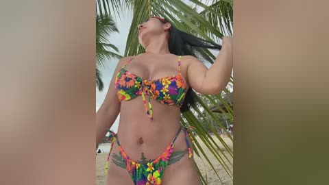 Media: Video of a curvy, light-skinned woman in a colorful, star-patterned bikini, standing beneath a palm tree on a sandy beach. Her long black hair cascades down, and she has a tattoo on her lower abdomen.