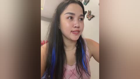 Media: A video of a young Asian woman with long, straight black hair featuring blue highlights, wearing a pink sleeveless top. She is indoors, partially obscured by two large, light brown objects. The background includes a white wall with butterfly stickers.