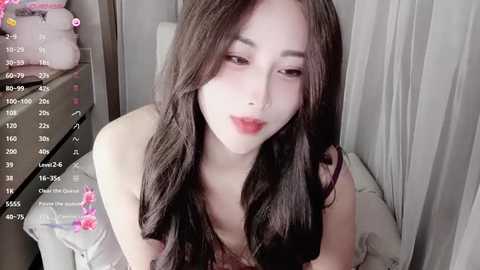 Media: Video of an East Asian woman with long, wavy black hair, fair skin, and red lipstick, wearing a light-colored top, sitting on a bed with white sheets.