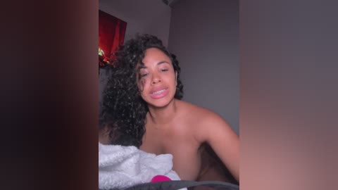 Media: A video of a topless Black woman with curly hair, wearing pink lipstick, smiling in a dimly lit room with a white blanket and red curtains in the background.