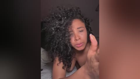 Media: Video of a topless Black woman with voluminous, curly hair, smiling, her hand touching her cheek. She has medium-brown skin and is indoors, likely in a bedroom.