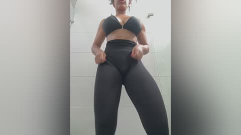 Media: Video of a dark-skinned woman with curly hair, wearing a black sports bra and high-waisted leggings, standing in a modern white-tiled bathroom. She's lifting her leggings, revealing her toned legs.