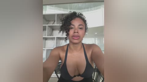 Media: Video of a Black woman with curly hair and medium skin tone, wearing a black bikini top, posing in a modern, well-lit living room with white bookshelves and glass railings in the background.