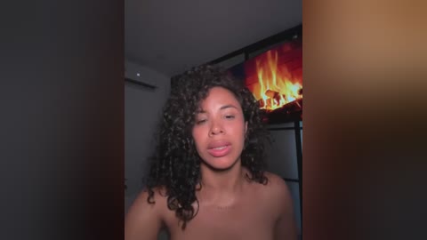 Media: Video of a topless Black woman with curly hair, eyes closed, in a dimly lit room with a burning fireplace in the background.