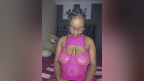 Media: Video of a dark-skinned woman with braids and glasses, wearing a revealing pink lace bodysuit, standing in a dimly lit bedroom with a teddy bear and framed art on the wall.