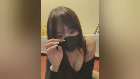 Media: Video of a young Asian woman with long black hair, wearing a black mask and off-shoulder top, kneeling on a red carpet in a room with beige walls and a bathtub.