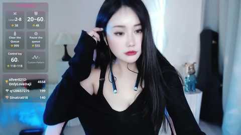 Media: Video of an Asian woman with long black hair, red lipstick, and black off-shoulder top, adjusting her hair. Background shows a dimly lit room with a lamp and a glass bottle.