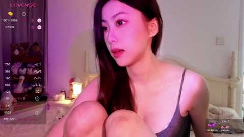 Media: A video of an Asian woman with long black hair, wearing a grey tank top, sitting on a bed in a dimly lit bedroom with pink lighting, watching a live stream on her phone.