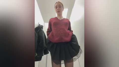 Media: Video of a slender, fair-skinned young woman with blonde hair in a tight bun, wearing a maroon sweater and a black pleated skirt, standing in a bright, modern dressing room with white walls and a grey floor.