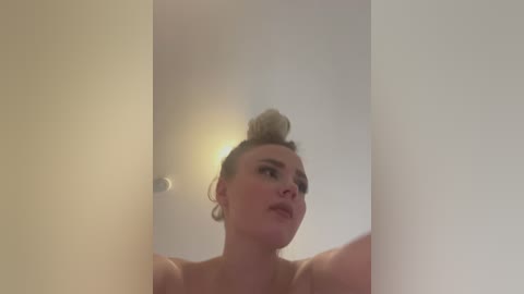 Media: Video of a light-skinned woman with blonde hair tied in a high bun, standing in a dimly lit room with white walls. She has a contemplative expression, wearing minimal makeup.