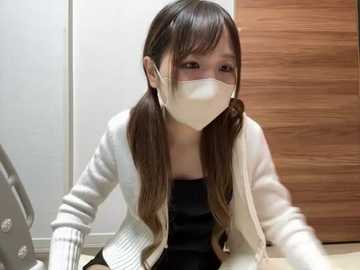 Media: Video of an Asian woman with long brown hair in pigtails, wearing a white mask, black top, and white cardigan, sitting on the floor in a modern, wooden-furnished room.