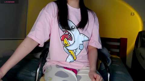 Media: Video of a person with long black hair, wearing a pink t-shirt featuring a cartoon shark with a yellow fork in its mouth. They sit on a blue-patterned bed, with a yellow wall and a dark wooden headboard in the background.