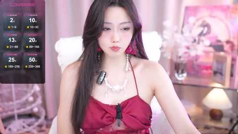 Media: Video of an East Asian woman with long black hair and fair skin, wearing a red lace lingerie top. Background features a cozy bedroom with floral decor and a nightstand lamp.