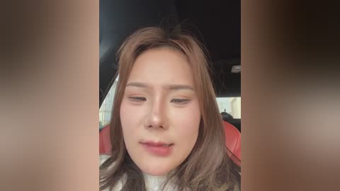 Media: Video of a young Asian woman with fair skin and shoulder-length brown hair, wearing makeup with subtle eyeshadow and lipstick. She is seated in a car with a red interior, looking serene.