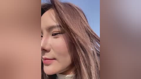 Media: A close-up video of an East Asian woman with long brown hair, fair skin, and delicate features, wearing subtle makeup, smiling slightly. The background is blurred, suggesting an outdoor setting.