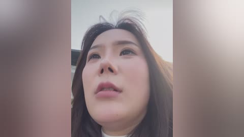 Media: Video of a young Asian woman with long, dark hair and fair skin, looking upwards with a neutral expression. Background is blurred with soft lighting, creating a serene and introspective atmosphere.