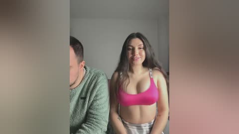 Video of a young woman with long brown hair, wearing a pink sports bra and high-waisted plaid shorts, standing next to a man in a green sweater, both indoors, with blurred background.