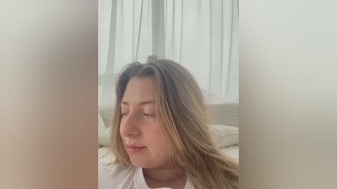 Media: A video of a young woman with long, wavy brown hair, fair skin, and light makeup, wearing a white shirt, gazing downward in a softly lit, minimalist bedroom with sheer curtains.