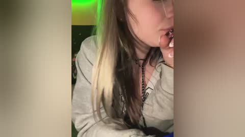 Media: Video of a young woman with long, multicolored hair, wearing a gray hoodie, biting her lower lip, with a green light in the background.