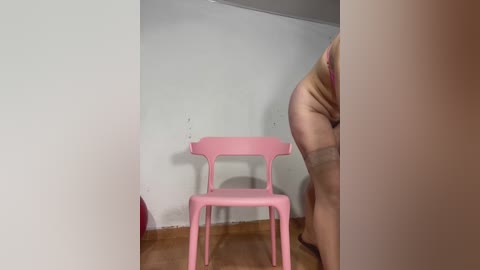 Media: Video of a nude person bending over, their backside partially visible, standing near a pink chair against a white wall in a sparsely furnished room.