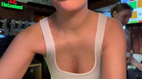 Media: A video of a woman in a white tank top with a plunging neckline, captured from the chest up in a dimly lit bar. The background features blurred bar fixtures and a Heineken sign.