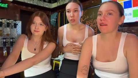 Media: Video of three young women, two with long hair, one with short hair, wearing white tank tops and black pants, posing in a bar with a colorful sign and shelves in the background.