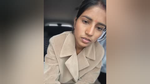 Media: Video of a South Asian woman with medium brown skin and straight black hair, wearing a beige trench coat, sitting in a car, with blurred background.