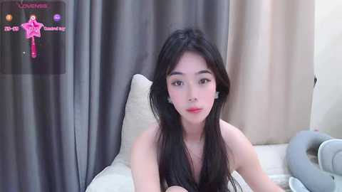 Media: Video of a young Asian woman with long black hair, fair skin, and red lipstick, lying on a white bed with gray curtains in the background.