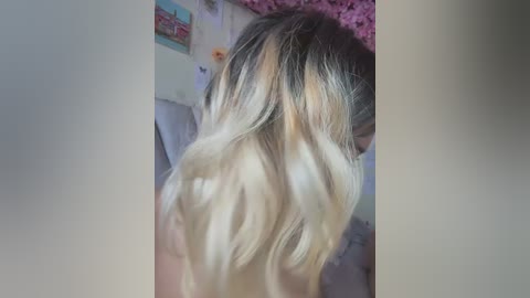 Media: A video of a nude, slender, blonde woman with long, wavy hair, viewed from behind. She is indoors, with a pink floral curtain in the background and a framed painting on the wall.