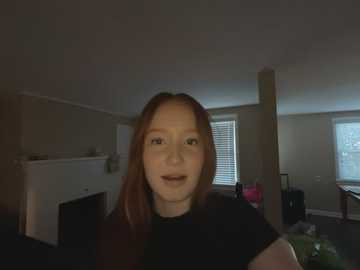 Media: A video of a young woman with light skin and straight brown hair, wearing a black top, standing in a dimly lit, sparsely furnished room with beige walls and blinds on windows.