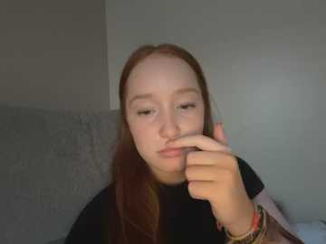 Media: Video of a young Caucasian woman with long red hair, wearing a black top, covering her mouth with her right hand. She has a neutral expression. Background is a gray sofa and a plain wall.