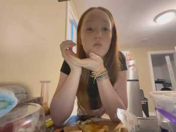 Media: A video of a young, fair-skinned girl with long, straight, reddish-brown hair, wearing a black T-shirt, eating a sandwich in a messy kitchen with cluttered countertops.