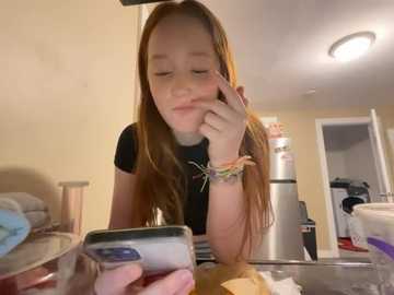 Media: A candid video of a young, fair-skinned girl with long, straight, reddish-brown hair, wearing a black t-shirt, resting her chin on her hand, smiling at a smartphone on a cluttered kitchen counter.