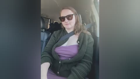 Media: Video of a light-skinned woman with shoulder-length brown hair, wearing dark sunglasses and a dark green cardigan over a purple dress, seated in a car with a black seatbelt.