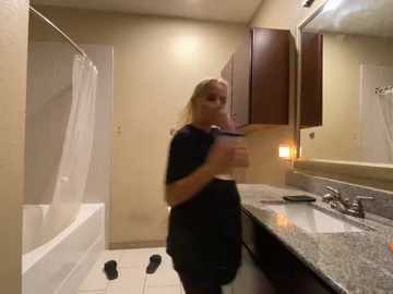 Media: Video of a blonde woman in a black shirt and pants, holding a baby, standing in a beige bathroom with a granite countertop, white sink, and dark wooden cabinets.