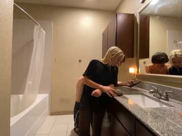 Media: Video of a blonde woman in a black t-shirt and jeans bending over a granite bathroom sink, next to a mirror and bathtub.