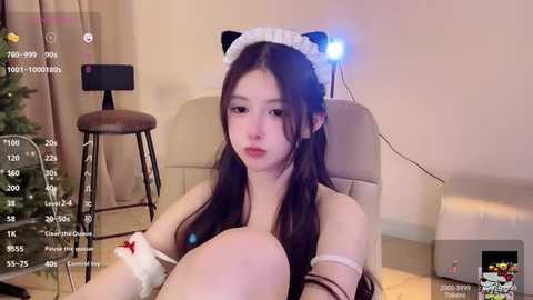 Media: A video of an East Asian woman with long black hair, dressed in a white maid headband and lingerie, sitting on a beige couch, in a room with a Christmas tree and a lamp.