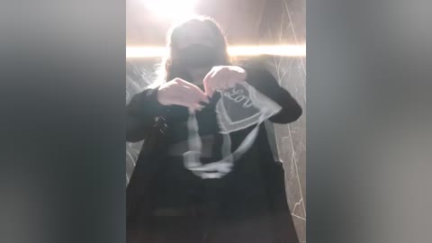 Media: Video of a person in a dimly lit room, wearing a black jacket and a white T-shirt with a graphic design, holding up a piece of paper with \"love\" written on it, creating a heart shape.