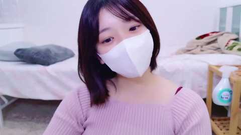 Media: Video of an East Asian woman with straight black hair, wearing a white face mask and a ribbed pink sweater, sitting on a bed in a cluttered room with a wooden shelf and a bottle of disinfectant.