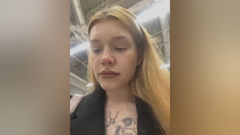 Media: Video of a fair-skinned, blonde young woman with closed eyes and a serene expression, wearing a black jacket. Her chest features a detailed tattoo of a bee. Background shows a dimly lit, industrial-style room with exposed pipes.