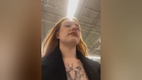 Media: Video of a young white woman with long, straight red hair and pale skin, wearing a black jacket, standing in an industrial warehouse with exposed ceiling and fluorescent lights. She has a tattoo on her chest.