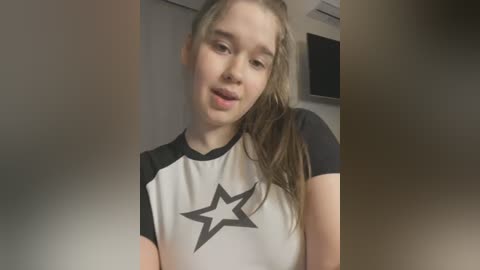 Media: Video of a young Caucasian woman with fair skin, straight blonde hair, and blue eyes, wearing a white and black star-printed athletic t-shirt, leaning slightly forward with a neutral expression. Background includes a beige wall and a black-framed TV.